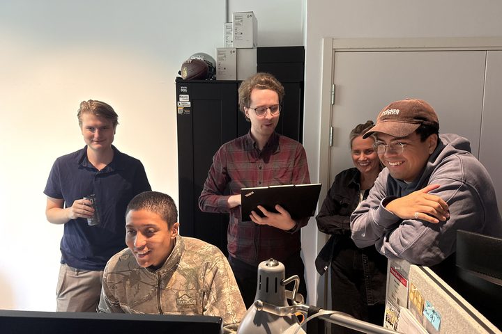 Five colleagues gathered around a desk screen, laughing during a build review.